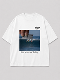 Wit oversized THE WAVE OF LIVING T-shirt van FAKESNIFF, print 'the wave of living': een hand houdt een scheermesje boven een golf waarop drie surfers rijden